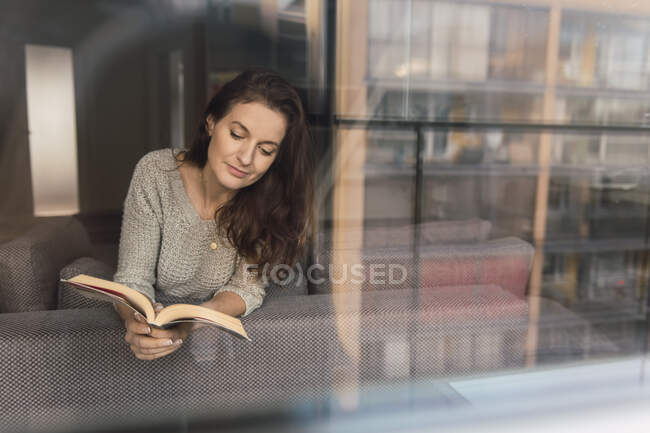 Adorável senhora adulta em roupa elegante sorrindo e olhando para longe enquanto deitado no sofá confortável com livro interessante — Fotografia de Stock