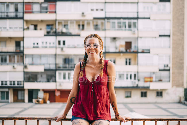 Pretty red-haired girl with braids and sunglasses leaning on railing against residential building — Stock Photo