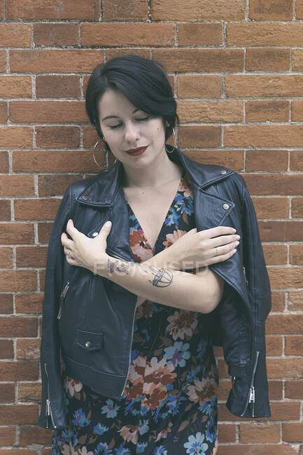 Young beautiful woman in leather jacket and dress with flower design standing near brick wall and looking at camera — Stock Photo
