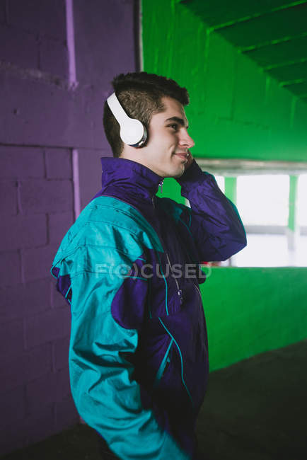 Улыбающийся молодой человек в спортивной одежде, слушающий музыку в наушниках у красочной стены — стоковое фото