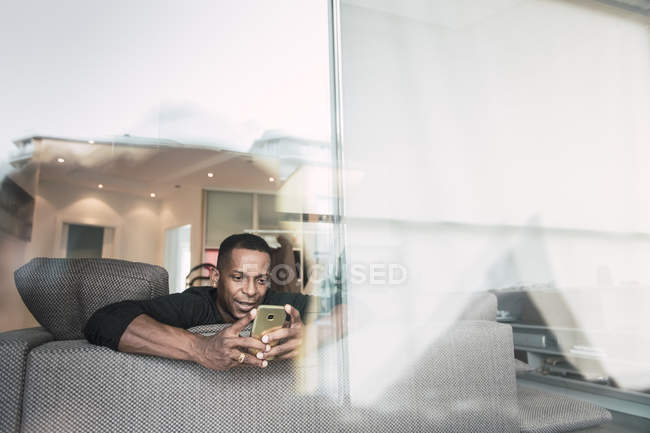 Hombre afroamericano usando teléfono inteligente en el sofá en casa - foto de stock