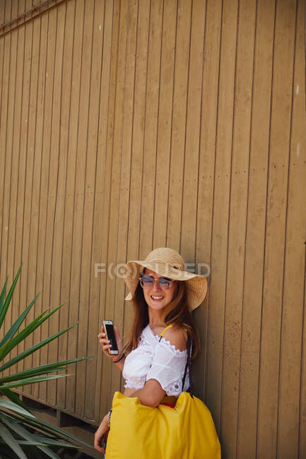 Donna sorridente in cappello di paglia con dispositivo? alla parete di legno — Foto stock