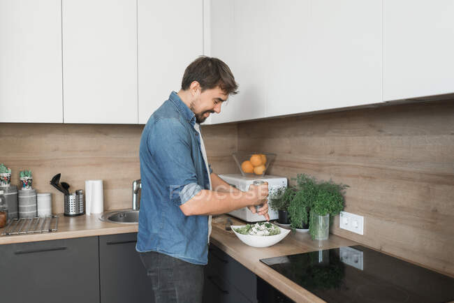 Vista lateral de chico joven guapo cortar lechuga fresca para deliciosa ensalada? mientras está de pie en la cocina con estilo - foto de stock