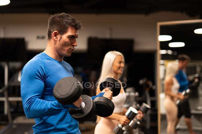 Homem de pulôver azul e mulher atraente em sportswear de pé no ginásio na frente do espelho e fazendo exercício com halteres — Fotografia de Stock