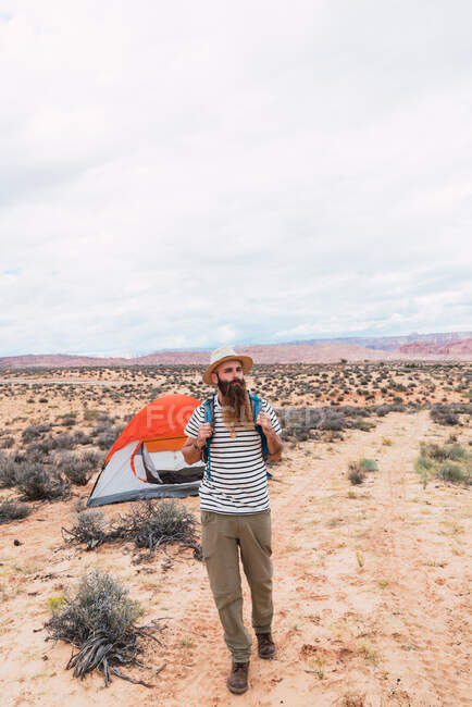 Schöner bärtiger Mann mit Rucksack schaut weg, während er an einem bewölkten Tag in der Wüste in Zeltnähe spaziert — Stockfoto
