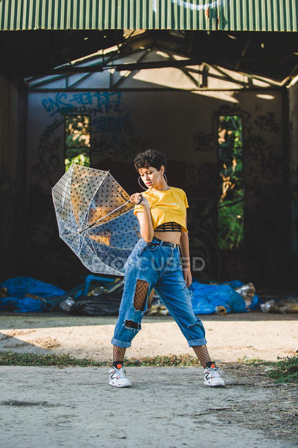 Mujer joven reflexiva en traje elegante posando con paraguas transparente mientras está de pie en la calle en el día soleado - foto de stock