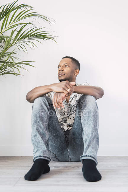 Плотный черный мужчина, сидящий на полу и отворачивающийся от белой стены с зеленым растением — стоковое фото