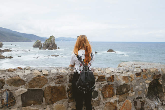 Visão traseira da jovem mulher com mochila em pé perto de parede de pedra velha e olhando para o belo mar na ilhota de Gatzelugatxe na Espanha — Fotografia de Stock