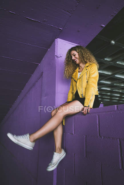 Стильная кудрявая блондинка в кроссовках и желтой куртке сидит на фиолетовой стене и улыбается — стоковое фото