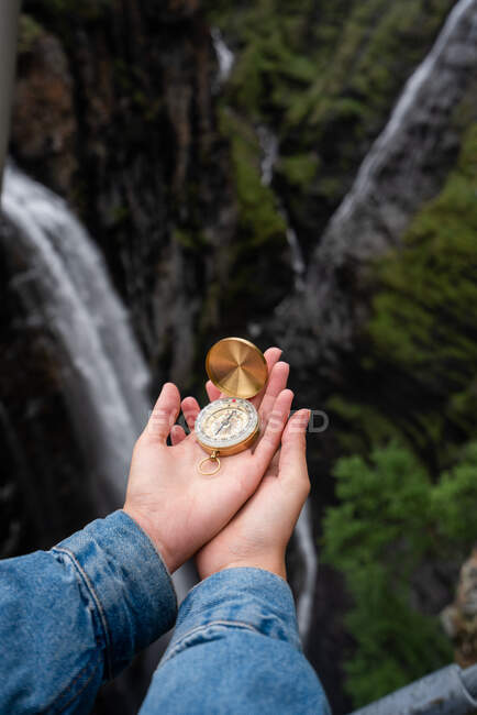 Crop view of hands of people holding golden compass on blurred background of waterfall in forest — Stock Photo