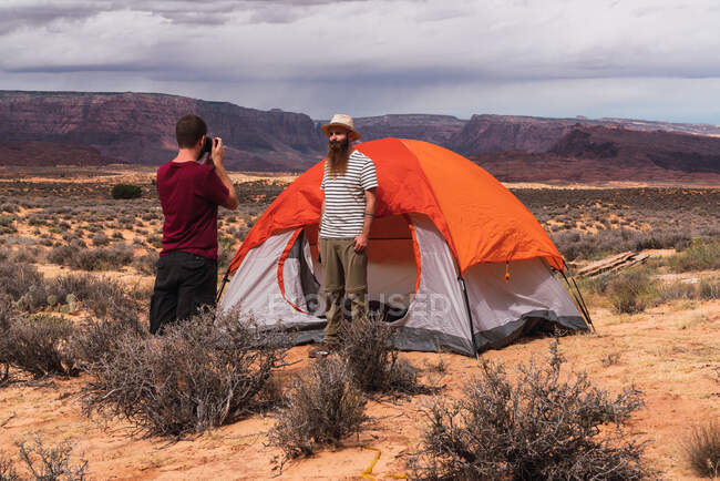 Due uomini belli in piedi vicino alla tenda e fare foto e video di incredibile deserto mentre viaggiano insieme — Foto stock