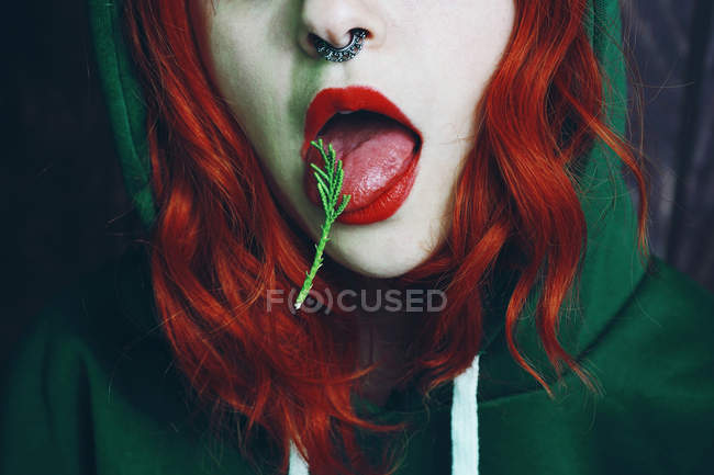 Gros plan de la femme rousse à capuche avec anneau dans le nez et bouche ouverte avec aiguille de conifères sur la langue — Photo de stock