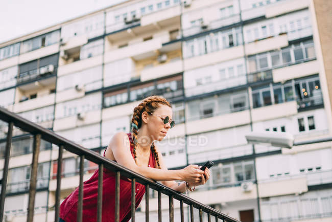 Red-haired young woman with braids using mobile phone while leaning on railing against residential building — Stock Photo
