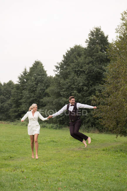Novio y novia adultos de moda en trajes elegantes tomados de la mano y saltando con emoción en el prado verde - foto de stock