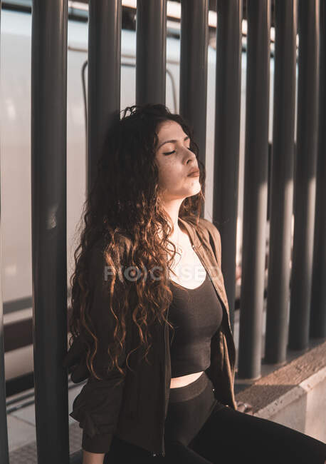 Beautiful young female in stylish outfit keeping eyes closed and enjoying bright sun while sitting near fence on city street — Stock Photo