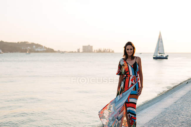 Содержательная женщина в длинном красочном платье ходит по булыжнику на закате против морской набережной — стоковое фото