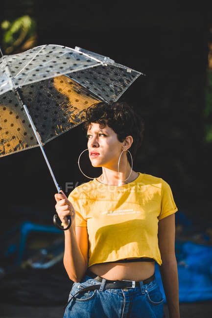 Jeune femme réfléchie avec parapluie transparent tout en se tenant dans la rue le jour ensoleillé — Photo de stock