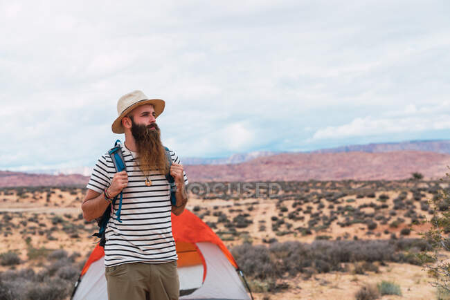 Handsome bearded man with backpack looking away while walking near tent on cloudy day in desert — Stock Photo