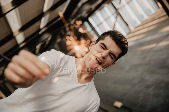Молодой красивый мужчина в белой футболке стоит внутри здания и держит горящий бенгальский свет в руке — стоковое фото
