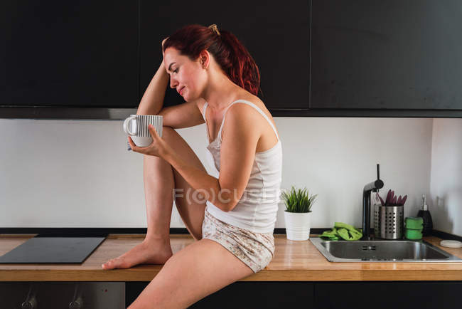Жінка сидить на кухонній стійці і п'є каву на кухні — стокове фото