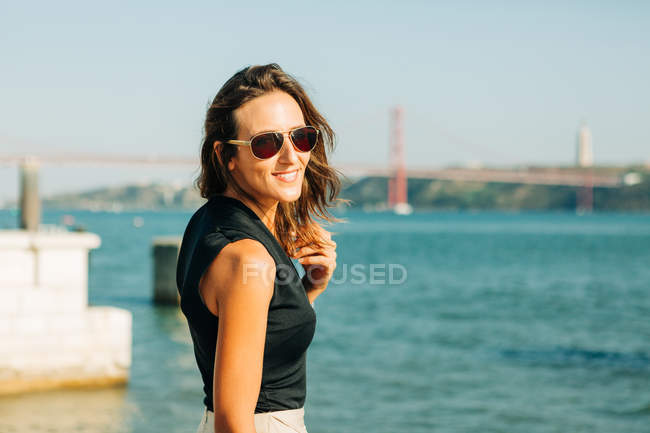 Jeune femme brune souriante debout sur le front de mer et regardant la caméra — Photo de stock