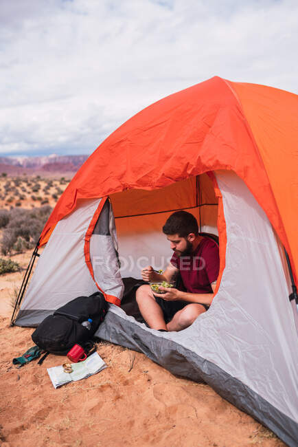 Crop man with bowl of fresh salad sitting at modern tent on camping area in desert — Stock Photo