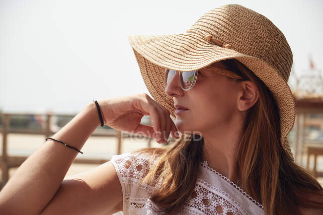 Attractive female with brown hair in sunglasses dressed in white shirt and straw hat on background with pier — Stock Photo