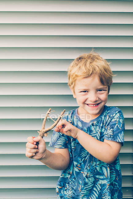 Petit garçon blond en t-shirt jouant avec fronde contre volets — Photo de stock