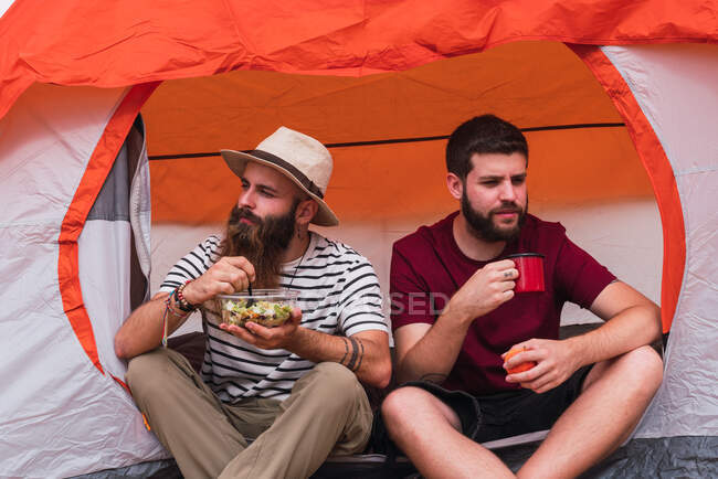 Два друга-мужчины улыбаются и сидят рядом с палаткой вместе и едят — стоковое фото