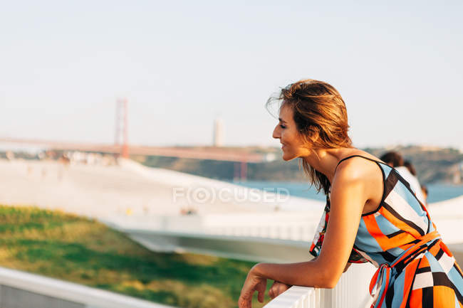 Mujer elegante sonriente en vestido apoyada en valla de puente y observando paisaje urbano a la luz del sol - foto de stock