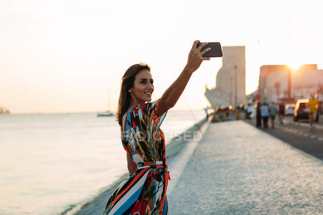 Conteúdo mulher em vestido usando telefone e tomando selfie à beira-mar ao pôr do sol — Fotografia de Stock