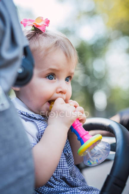 Lustiges Baby-Mädchen beißt Plastikspielzeug und schaut weg, während es im Kinderwagen auf verschwommenem Hintergrund des Parks sitzt — Stockfoto