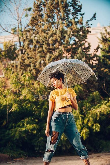 Attractive young woman in stylish casual outfit looking at camera while holding umbrella and standing on street near bushes — Stock Photo