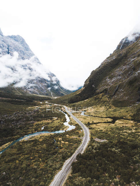 Spektakulärer Drohnenblick auf schmale Straße und Fluss, die durch das Tal inmitten majestätischer Berge führen, an bewölkten Tagen im Fiordland National Park in Neuseeland — Stockfoto