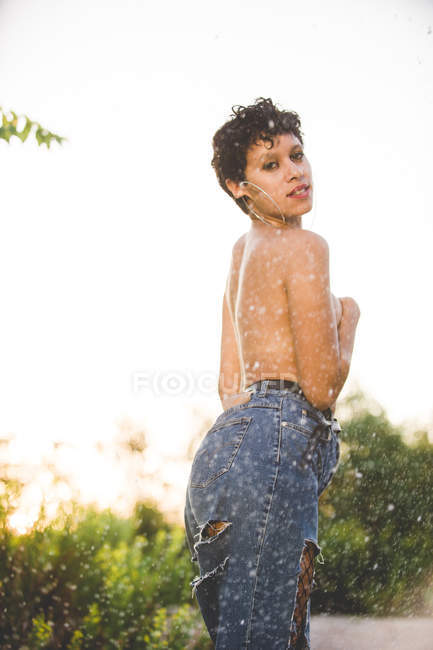 Уверенная в себе провокационная женщина в джинсах в джинсах, стоящая топлесс, прикрывающая грудь и смотрящая на камеру в природе — стоковое фото
