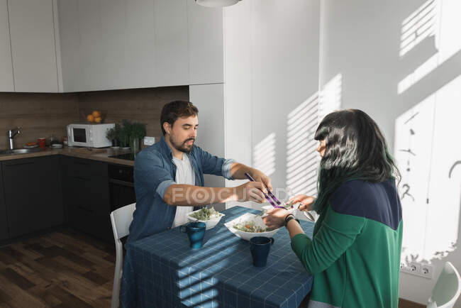Side view of young man and woman sitting at table in stylish apartment and enjoying fresh vegetable salad — Stock Photo