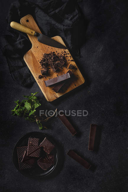 Chocolate pieces and chunks with mint on wooden board on black background — Stock Photo