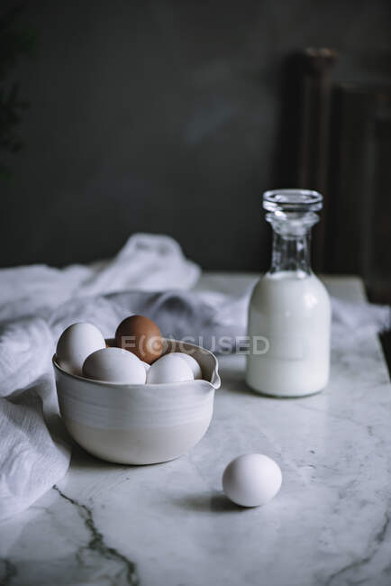 Cuenco de huevos de pollo y botella de lácteos frescos de pie en la mesa de mármol en la cocina - foto de stock