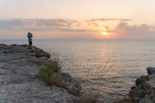 Вид сбоку неузнаваемого мужчины, стоящего на грубой скале и фотографирующего великолепный закат над морем в Тюленово, Болгария — стоковое фото