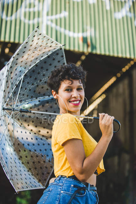 Portrait de jeune femme avec parapluie transparent souriant et regardant la caméra tout en se tenant debout dans la rue — Photo de stock