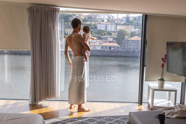 Вид ззаду чоловіка, загорнутий в рушник після душу, що стоїть з дитиною на руках біля панорамного вікна — стокове фото
