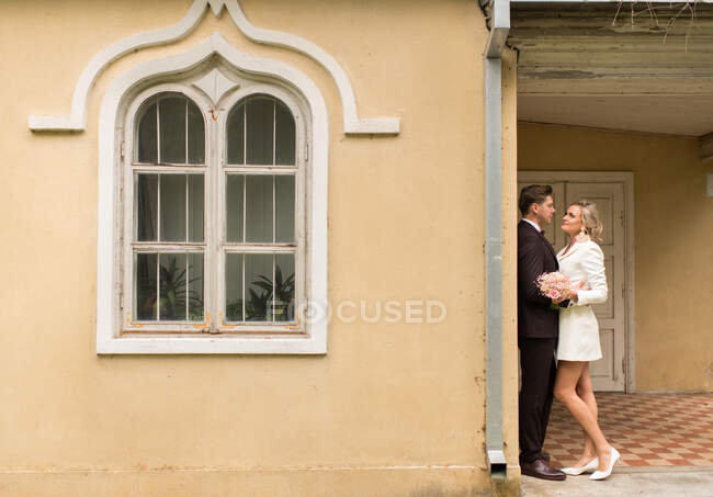Vista laterale di eleganti sposi che si abbracciano vicino a una piccola vecchia casa nel giardino verde — Foto stock