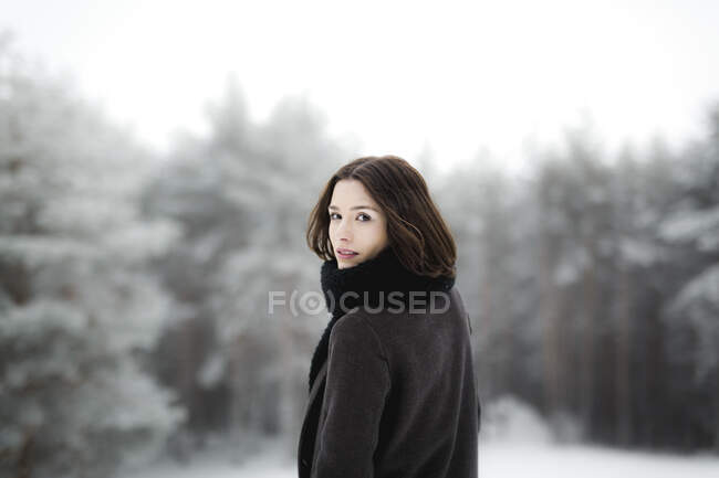 Красива молода жінка в стильному одязі дивиться у далечінь, стоячи біля дерева, вкритого снігом в холодний день у прекрасній сільській місцевості. — стокове фото