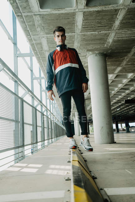 Jeune homme beau en veste de sport marchant sur rail de fer dans un bâtiment spacieux avec des piliers — Photo de stock
