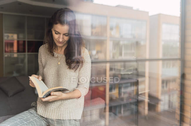 Mulher adulta com livro de pé perto do sofá e da janela — Fotografia de Stock