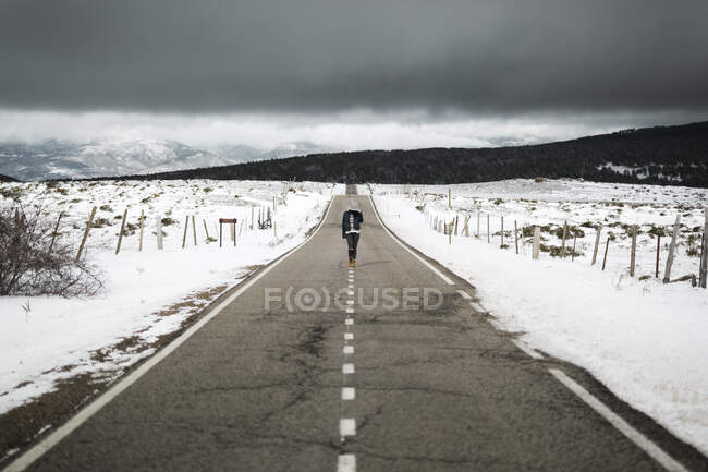 Vue arrière de la jeune personne en tenue élégante marchant au milieu de la route asphaltée par une journée d'hiver nuageuse dans une belle campagne — Photo de stock