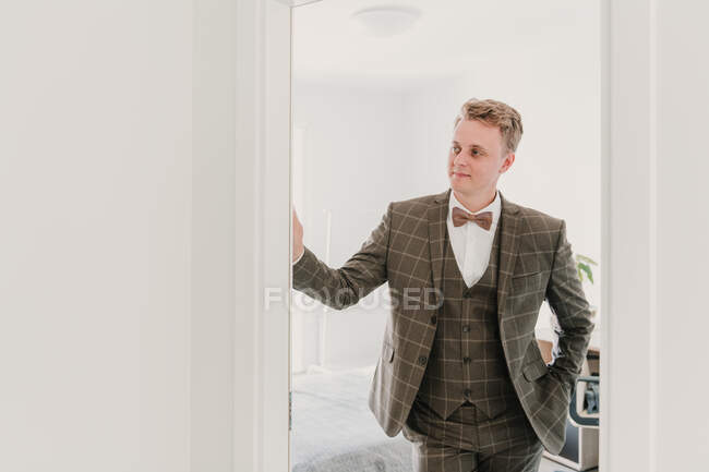 Guapo joven en elegante traje de novia sonriendo y mirando hacia otro lado mientras está de pie en la puerta de la habitación con estilo - foto de stock