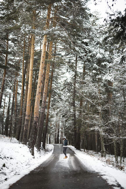 Вид сбоку молодого человека в стильной одежде, стоящего на асфальтовой лесной дороге в величественный зимний день — стоковое фото