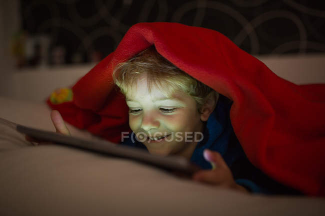 Sorrindo menino assistindo desenhos animados com tablet digital na cama sob cobertor — Fotografia de Stock