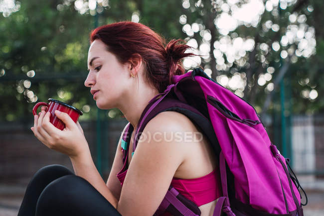 Junge Sportlerin mit Rucksack sitzt draußen und trinkt Kaffee — Stockfoto
